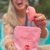 A smiling woman by a pool holding the pink stimulator above a matching pink pouch with playful embroidered text.