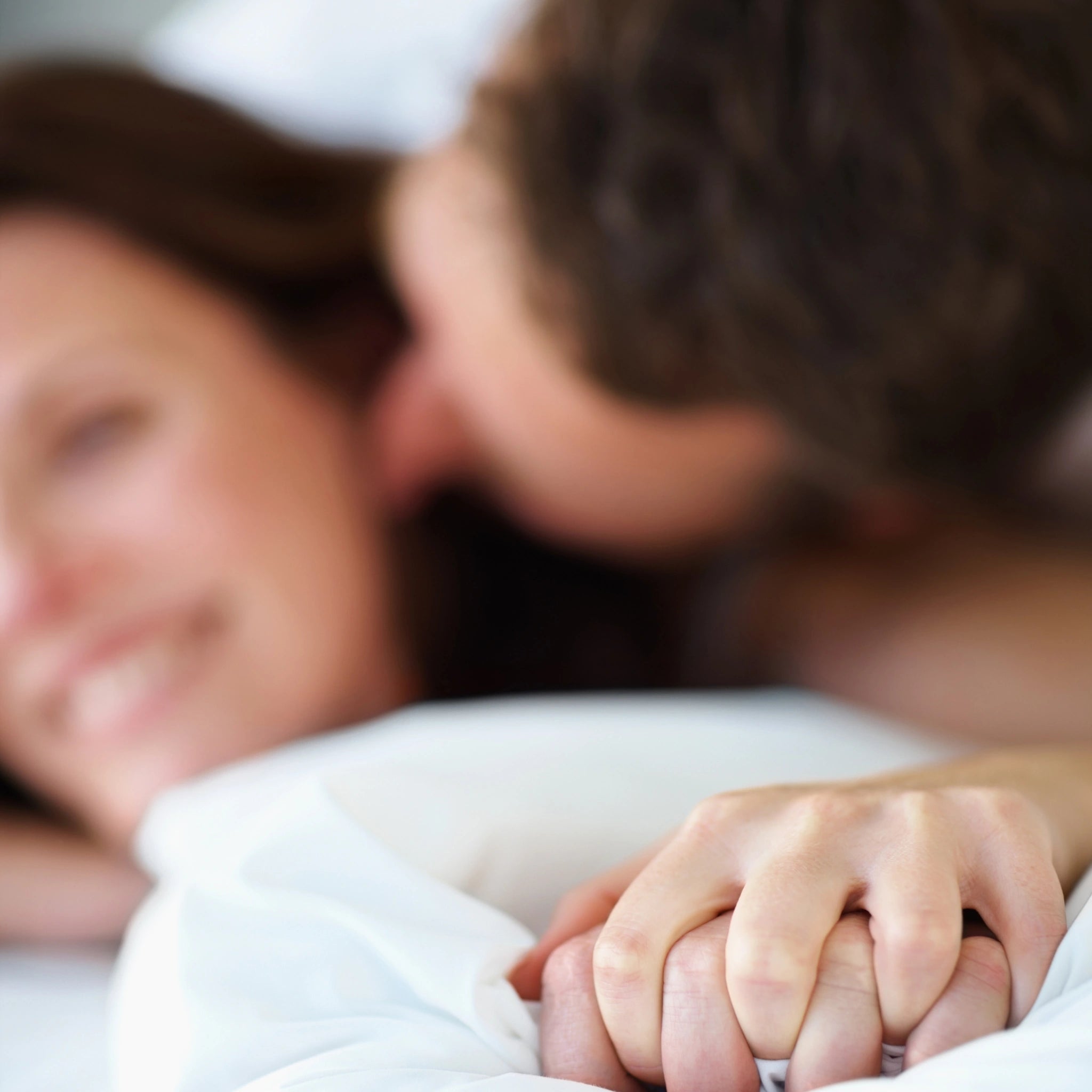 Close-up of a couple in bed, woman smiling and man kissing her, with their hands clasped in the foreground.