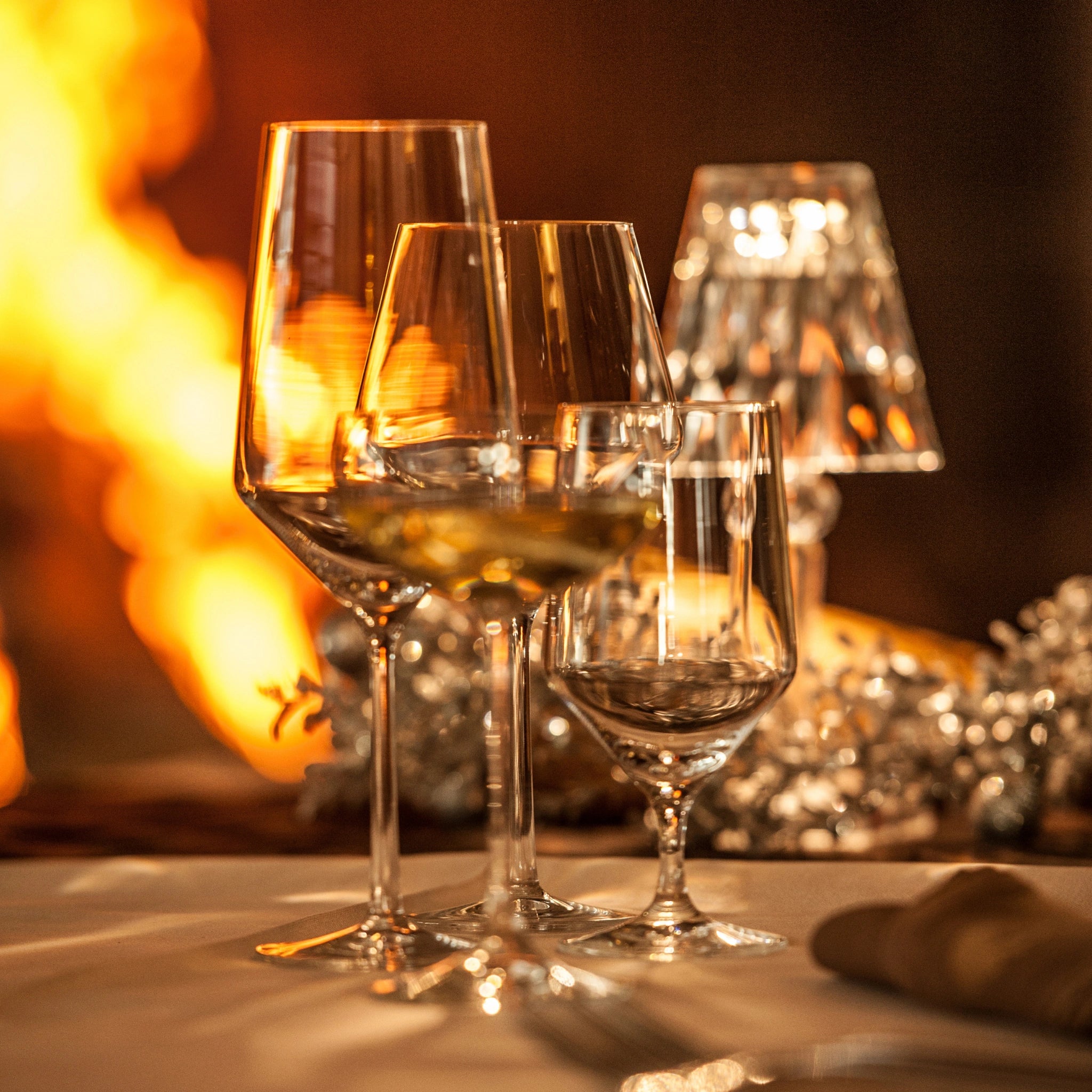 Elegant wine glasses on a table with a warm glowing fireplace and soft lights creating a cozy romantic dinner setting.