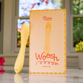 Yellow wand massager standing next to its bright packaging that reads “Woosh” by Oboo, placed on a sunny windowsill.