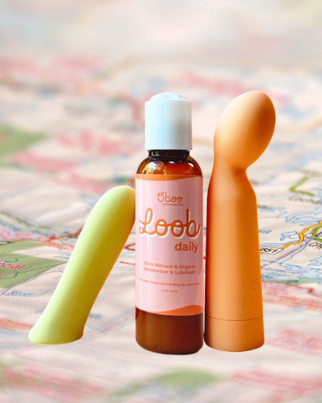 A bottle of Oboo Loob Daily lubricant is placed between two colorful massagers on a soft, blurred background of a map.