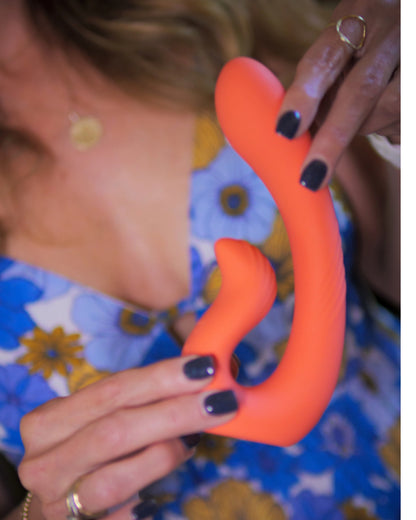 A close-up of a woman in a floral dress holding a red dual-action vibrator with one hand above and one below.