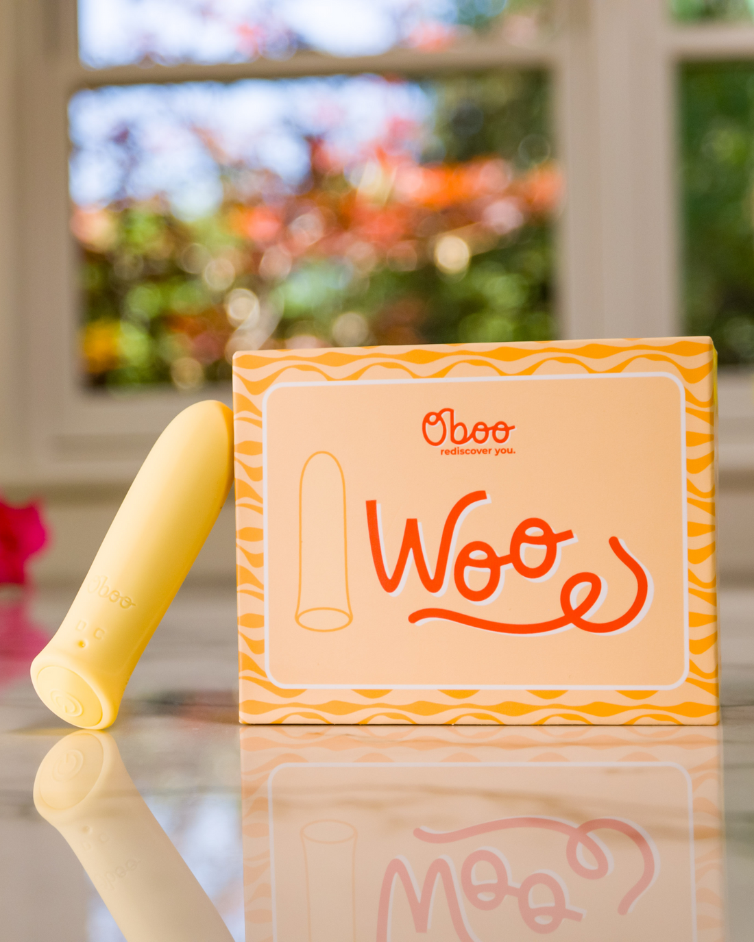 A yellow Woo massager stands beside its matching Oboo product box on a reflective surface, with soft natural light in background.